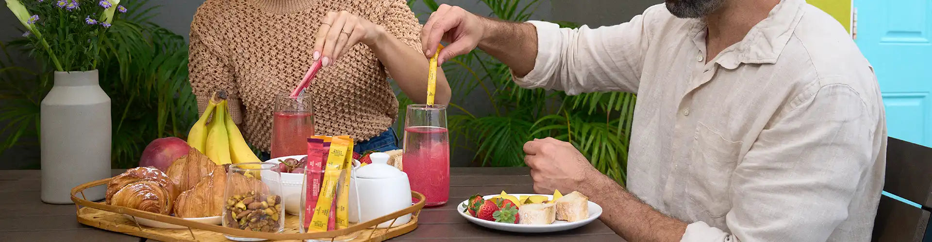 A couple enjoys a breakfast of pastries, fruit, and drinks on an outdoor table with a plant in the background.