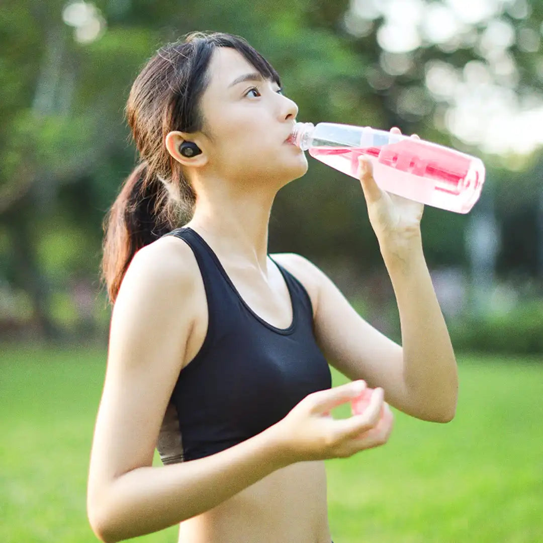 Woman in workout gear drinking water from a bottle outdoors, with greenery in the background.