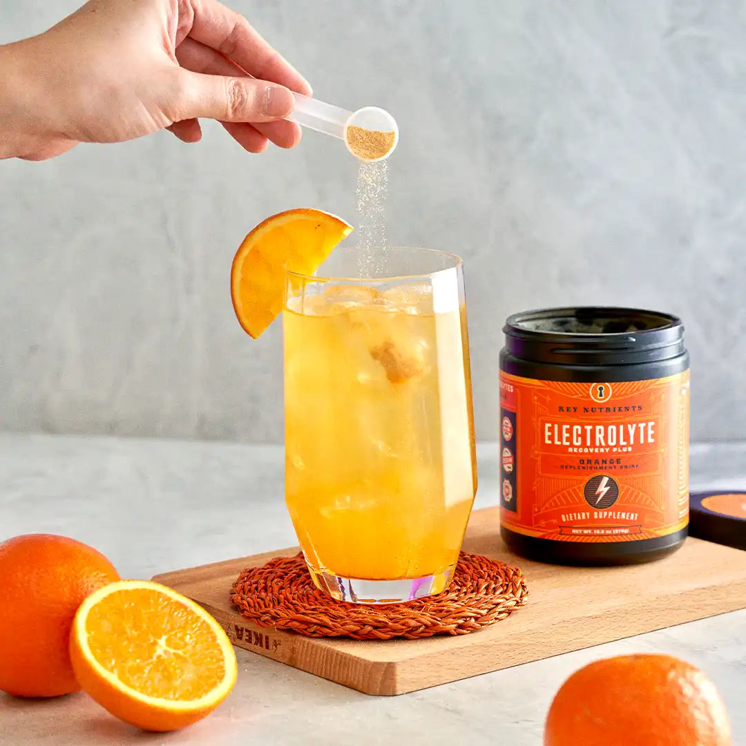 Hand adding powder to orange drink in glass, surrounded by oranges and a jar of electrolyte mix.