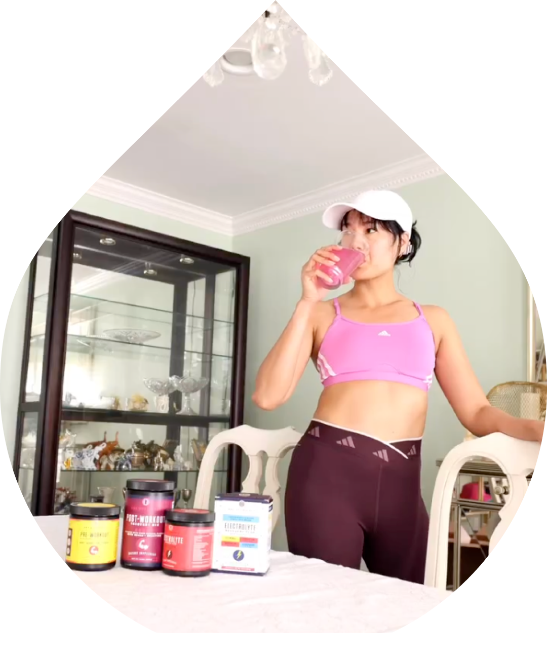 Person in workout attire drinking a pink beverage, with supplement containers on the table.