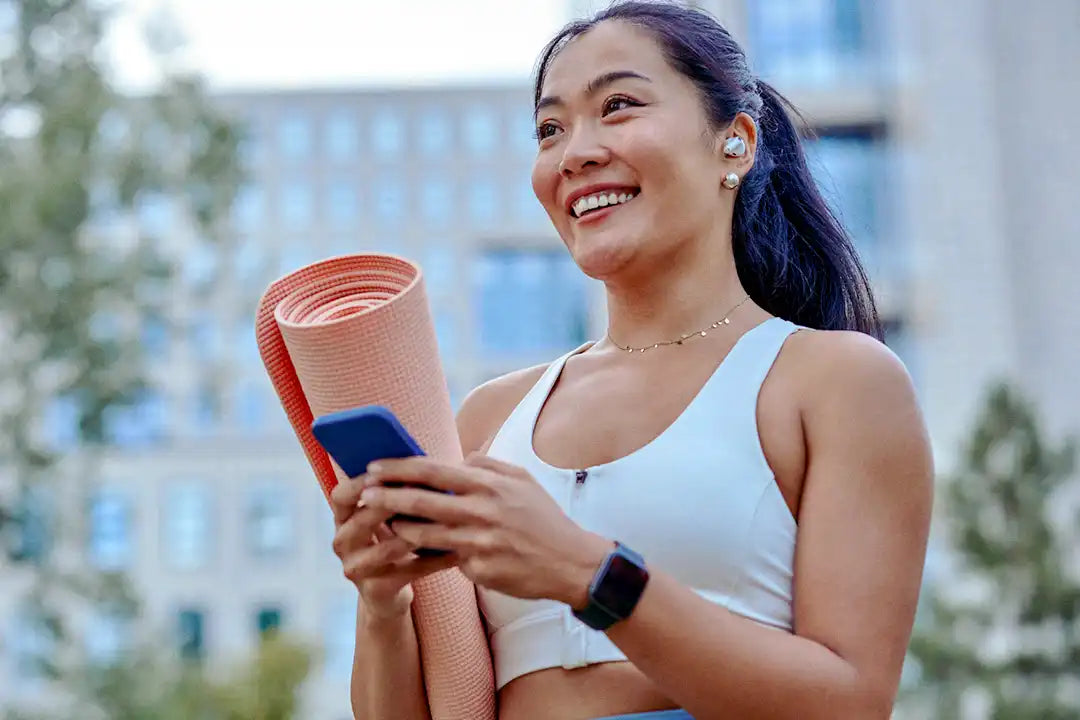 Smiling woman holding a yoga mat and phone, wearing earbuds, outdoors in an urban setting.