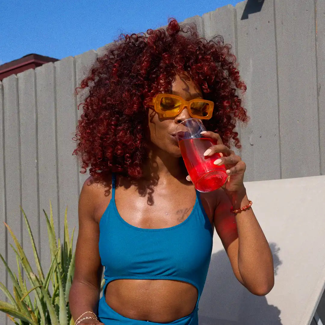Person in blue swimsuit drinking a red beverage outdoors with sunglasses.