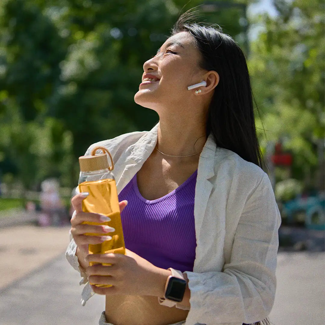 Woman smiling outdoors, holding a bottle, wearing earbuds and a smartwatch, with trees in the background.