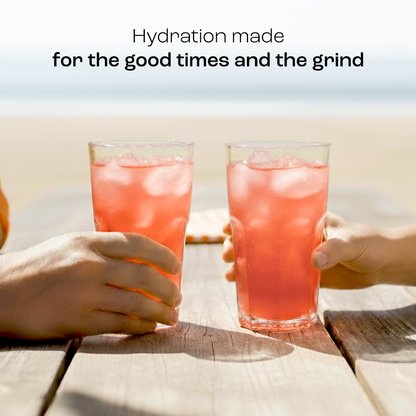 Two hands clink glasses of Electrolyte Recovery Plus Powder (Sugar-Free) by the beach, text above hydration.