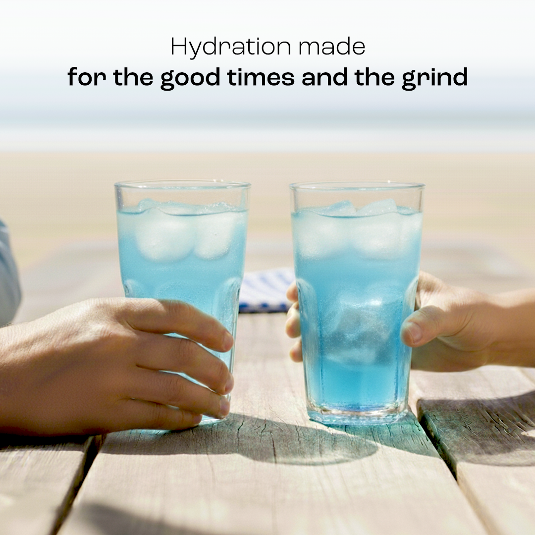 Two hands hold glasses of Electrolyte Recovery Plus Powder (Sugar-Free) by the beach, hydration text above.