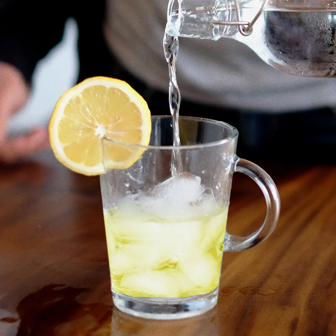 A glass mug with lemon slice and ice, being filled with water from a pitcher, on a wooden table.